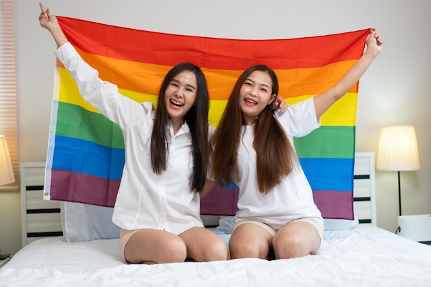 Ritratti di una felice coppia lesbica asiatica seduta con la bandiera dell'arcobaleno seduta accanto al letto