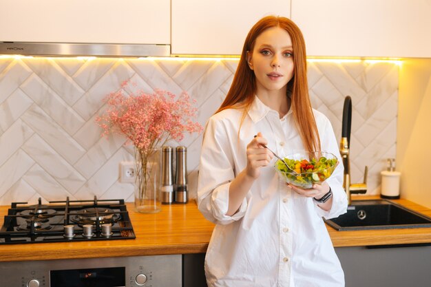 Ritratto a colpo medio di giovane donna attraente della testarossa che mangia insalata vegetariana fresca che gode del fresco