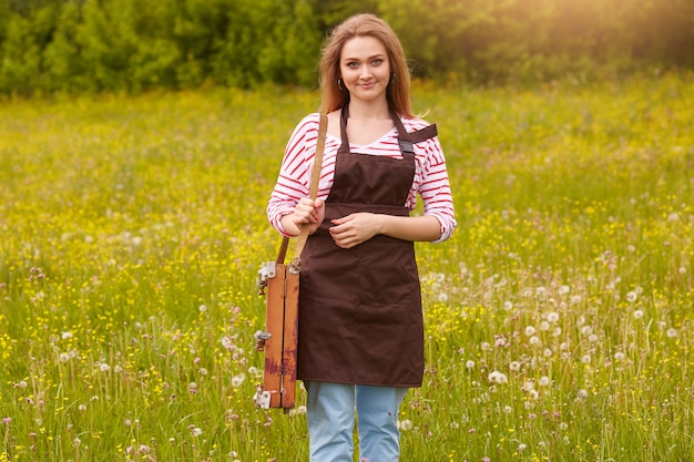 Ritratto a figura intera di bella, piacevole dall'aspetto signora con capelli chiari, indossa una camicia bianca con strisce rosse, pantaloni blu e grembiule marrone, in posa con attrezzature da disegno all'aria aperta