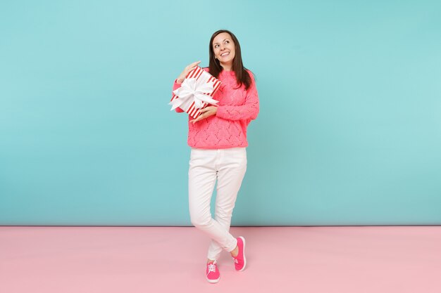 Ritratto a figura intera di donna in maglione rosa lavorato a maglia, pantaloni bianchi in posa con scatola regalo