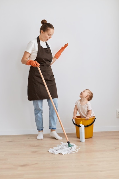 Ritratto a figura intera di donna scioccata che indossa una maglietta bianca e un grembiule in posa con il suo bambino che pulisce il suo appartamento tenendo il mocio guardando il bambino nel secchio con la bocca aperta