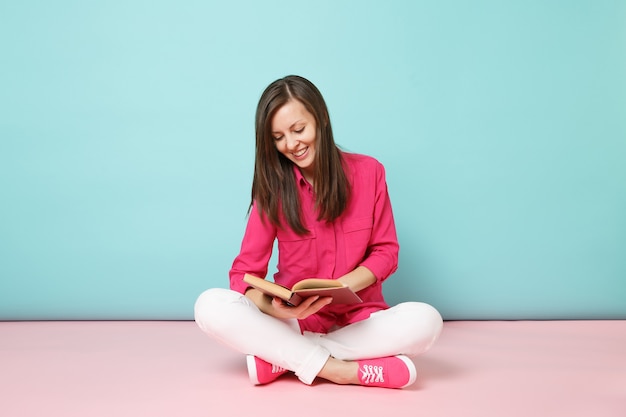 Ritratto a figura intera di donna sorridente in camicetta rosa, pantaloni bianchi seduti sul pavimento, leggere un libro