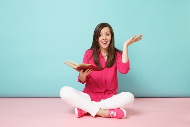 Ritratto a figura intera di donna sorridente in camicetta rosa, pantaloni bianchi seduti sul pavimento, leggere un libro