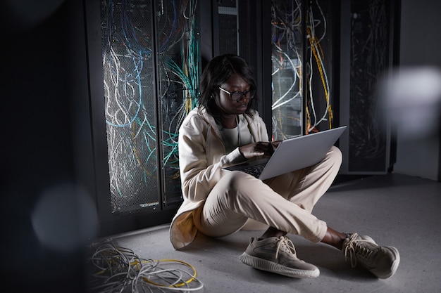 Ritratto a figura intera di giovane donna afro-americana seduta sul pavimento nella sala server e utilizzando il laptop mentre si lavora con il supercomputer al data center