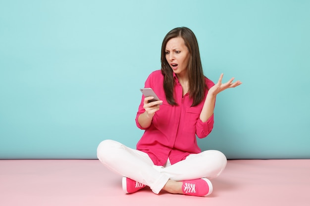 Ritratto a figura intera di giovane donna divertente in camicia rosa, pantaloni bianchi seduti sul pavimento tenere il cellulare