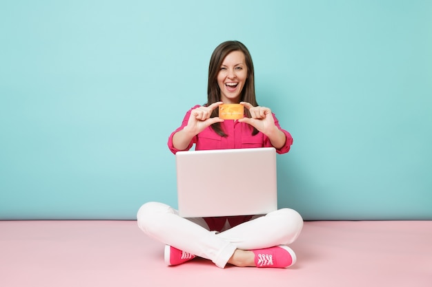 Ritratto a figura intera di giovane donna in camicia rosa, pantaloni bianchi seduta sul pavimento, tenere in mano un pc con carta di credito credit