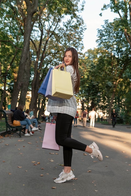 Ritratto a figura intera di giovane ragazza bruna con borse della spesa di carta a piedi nel parco Cliente felice Cornice verticale