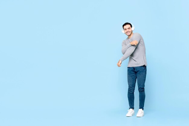 Ritratto a figura intera di giovane uomo caucasico bello sorridente che indossa la cuffia ascoltando musica mentre balla in uno sfondo blu studio isolato