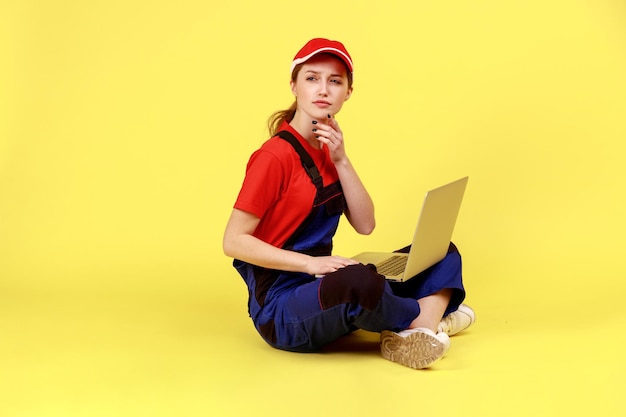 Ritratto a figura intera di lavoratrice pensierosa seduta sul pavimento con le gambe incrociate e lavorando su un taccuino, guardando da parte, pensando, indossando tute. Studio indoor girato isolato su sfondo giallo.