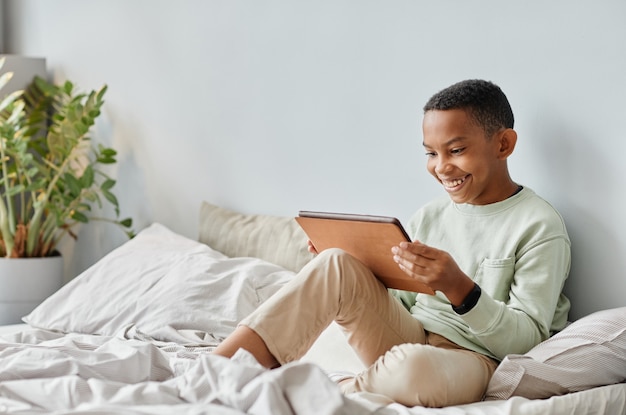 Ritratto a figura intera di un adolescente afroamericano sorridente che utilizza tablet mentre è seduto sul letto in un'accoglie...