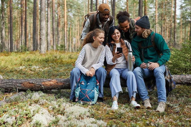 Ritratto a figura intera di un gruppo eterogeneo di amici seduti nella foresta di accesso e che utilizzano lo smartphone mentre...