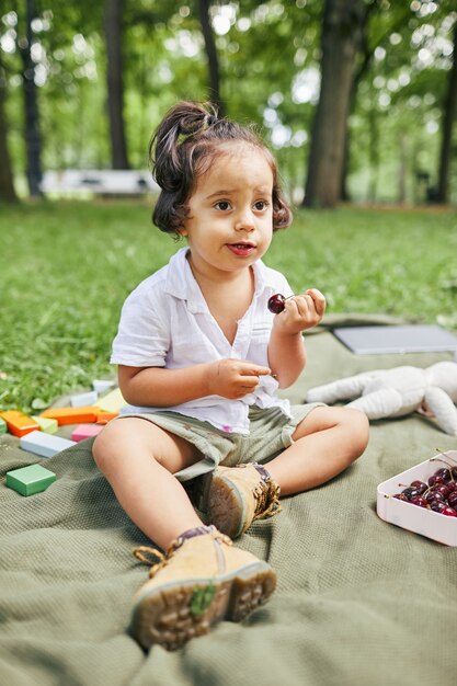 Ritratto a figura intera di un ragazzino carino seduto sull'erba verde nel parco e che mangia bacche mentre...