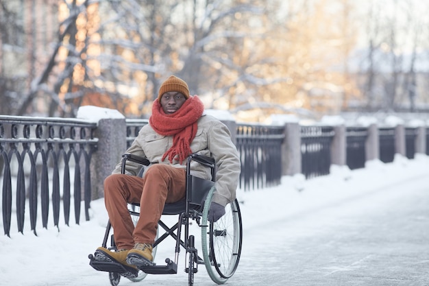 Ritratto a figura intera di un uomo afroamericano che utilizza una sedia a rotelle all'aperto in inverno e guarda la macchina fotografica, copia spazio