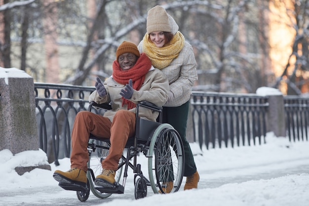 Ritratto a figura intera di un uomo afroamericano che utilizza una sedia a rotelle che si diverte all'aperto in inverno con una giovane donna sorridente che assiste, copia spazio