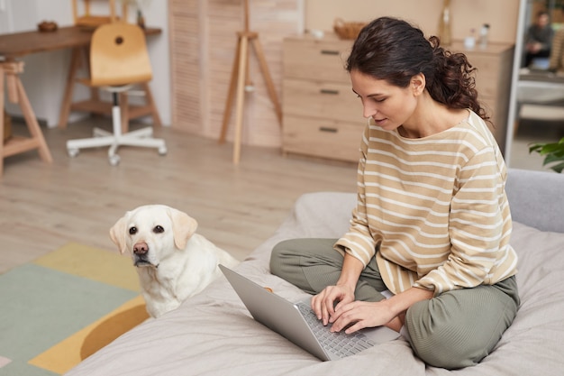 Ritratto a figura intera di una giovane donna sorridente che utilizza il computer portatile sul letto con un cane da compagnia in un accogliente interno domestico, copia spazio