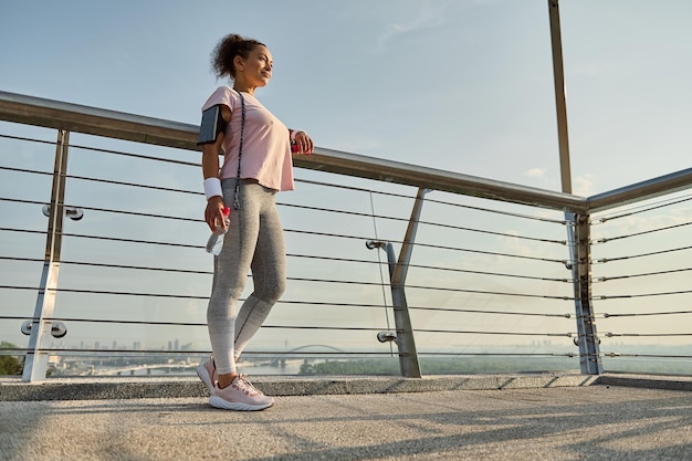 Ritratto a figura intera di una sportiva, atleta femminile con una corda per saltare, in piedi sul moderno ponte di vetro della città, rilassante dopo l'allenamento cardio all'aperto. Sport, fitness, concetto di stile di vita attivo