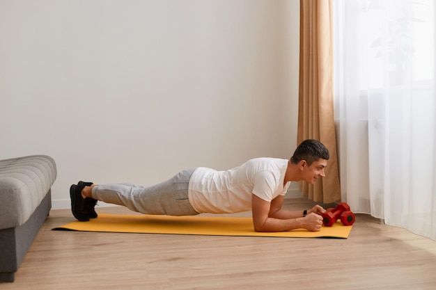 Ritratto a figura intera di uomo che indossa abbigliamento sportivo facendo esercizi sportivi a casa praticando yoga facendo esercizio Phalakasana sui gomiti allenando i muscoli facendo posa della plancia alta