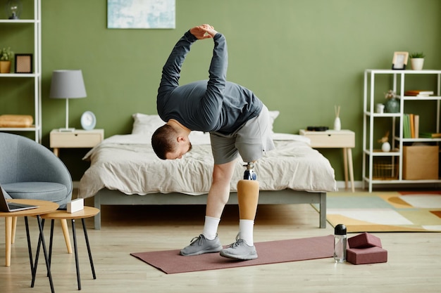 Ritratto a figura intera di uomo con gamba protesica che si allena a casa e fa esercizi di stretching c