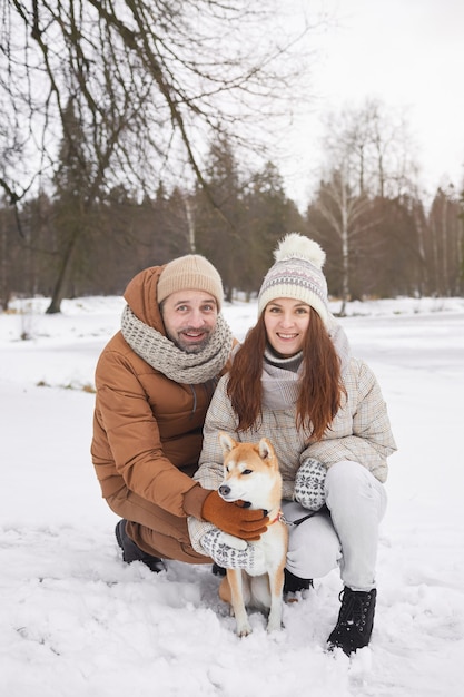 Ritratto a figura intera verticale di una coppia adulta felice che posa con il cane all'aperto e guarda la telecamera mentre si gode una passeggiata invernale insieme