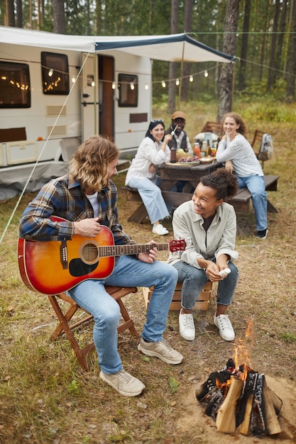 Ritratto a figura intera verticale di una giovane coppia che suona la chitarra mentre si accampa con gli amici nella foresta