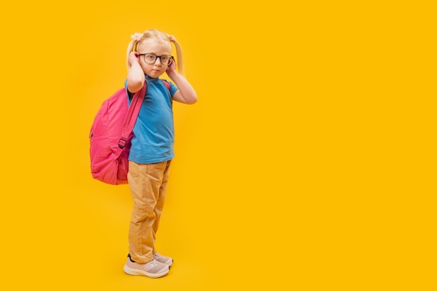 Ritratto a lunghezza intera di una bambina bionda seria che indossa uno zaino per lo sfondo giallo della scuola Mockup dello spazio di copia