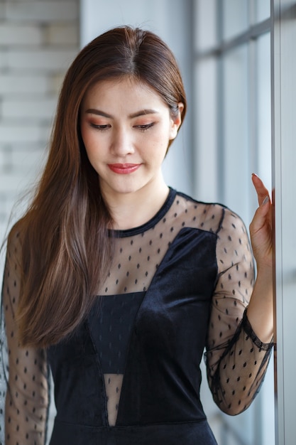 Ritratto a metà lunghezza di una donna asiatica bruna affascinante carina, etnia Lao, in elegante abito nero in piedi e posa con posa amichevole positiva e sicura di sé vicino alla finestra di vetro.