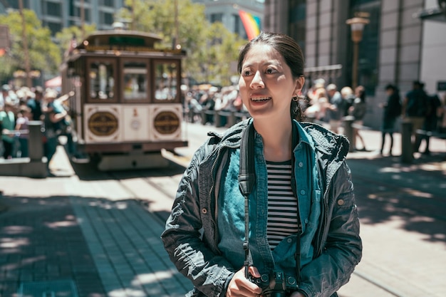 Ritratto a mezzo busto di una turista allegra che tiene la macchina fotografica e ride durante il giorno per passeggiare e visitare la città di san francisco. famosa funivia sullo sfondo. la signora sorridente gode del chiarore del sole