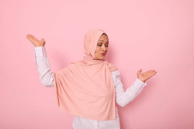 Ritratto a vita di bella misteriosa donna musulmana che indossa un velo hijab, mostrando qualcosa sui suoi palmi isolato su sfondo rosa colorato con copia spazio per la pubblicità o la promozione