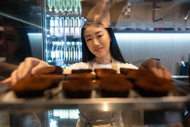 Ritratto a vita di un soddisfatto operaio di un caffè asiatico appoggiato sullo scaffale della vetrina per pasticceria