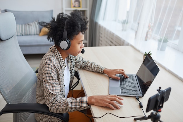 Ritratto ad alto angolo di un adolescente afro-americano che indossa l'auricolare utilizzando il laptop durante lo streaming di videogiochi a casa, concetto di giovane giocatore o blogger, spazio di copia