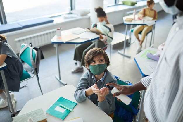 Ritratto ad alto angolo di un ragazzo che indossa la maschera e si disinfetta le mani nell'aula scolastica, copia spazio