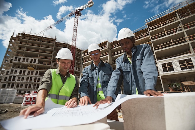 Ritratto ad angolo basso di tre lavoratori che discutono le planimetrie nello spazio della copia del cantiere