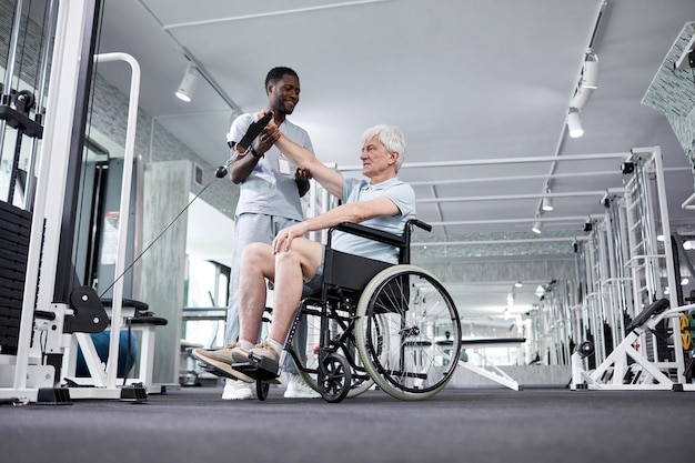 Ritratto ad angolo basso di un uomo anziano che usa una sedia a rotelle in palestra con un terapista riabilitativo che assiste il poliziotto