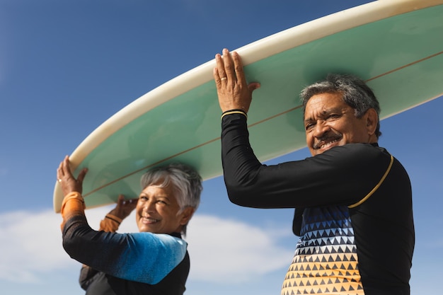 Ritratto ad angolo basso di una coppia senior multirazziale felice che trasporta la tavola da surf sopra la testa contro il cielo