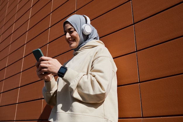 Ritratto ad angolo basso di una donna mediorientale contemporanea che utilizza lo smartphone in città illuminata dalla luce del sole e indossa le cuffie, copia spazio