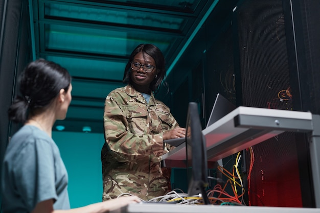 Ritratto ad angolo basso di una giovane donna afro-americana che indossa l'uniforme militare che utilizza il computer durante la configurazione della rete nella sala server