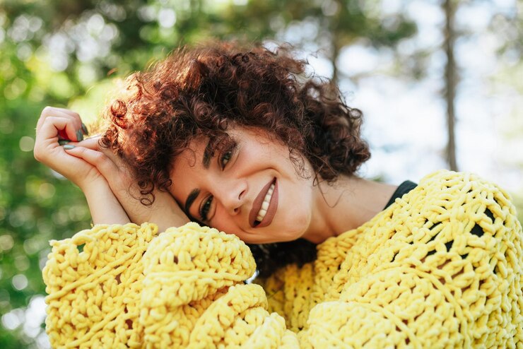 Ritratto all'aperto del primo piano di giovane donna attraente con i capelli ricci e sorridente La femmina ha un'espressione positiva che indossa un maglione giallo lavorato a maglia e posa contro lo sfondo della natura Stile di vita delle persone