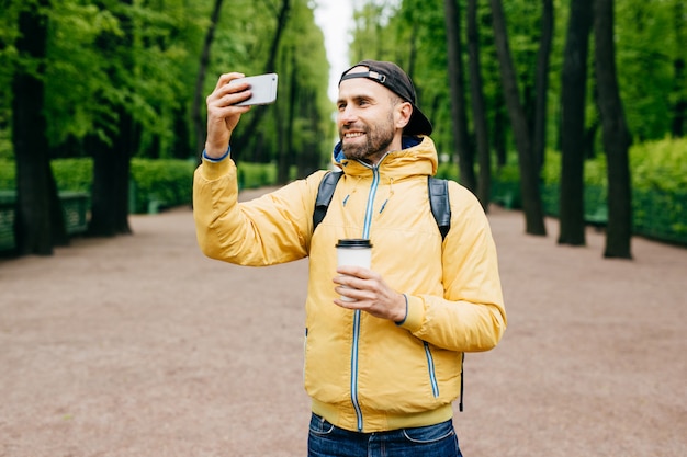 Ritratto all'aperto dell'uomo alla moda con barba di barba che indossa giacca a vento gialla e che tiene zaino e caffè da asporto facendo selfie con il suo cellulare