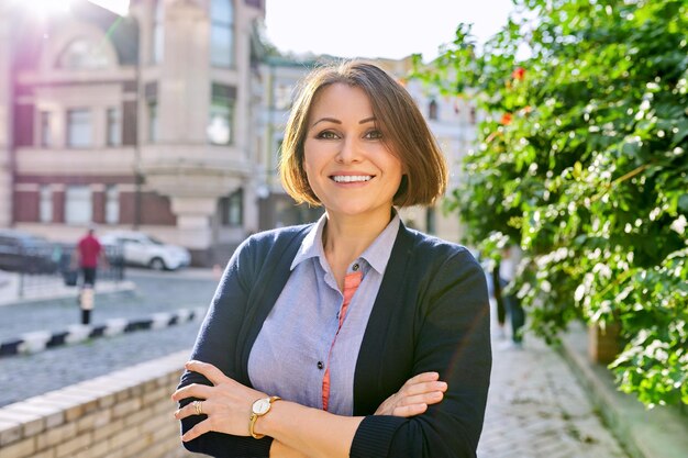 Ritratto all'aperto di donna sorridente di affari maturi con le braccia incrociate. Donna di mezza età positiva di successo che esamina la macchina fotografica, priorità bassa della via della città di tramonto