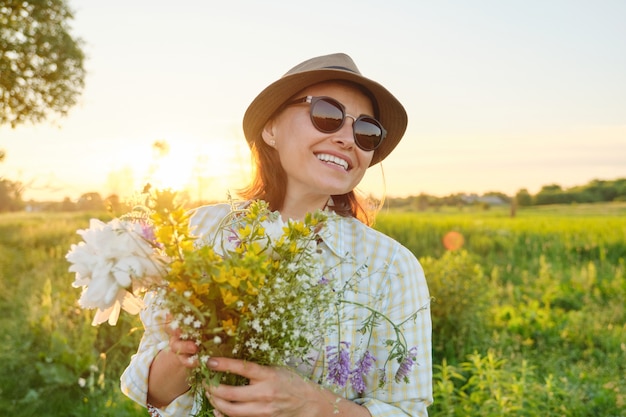 Ritratto all'aperto di donna sorridente felice matura con fiori primaverili, femmina in cappello da sole