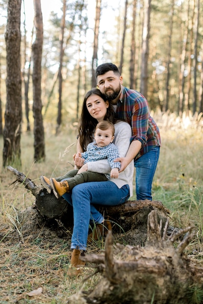 Ritratto all'aperto di famiglia caucasica alla moda felice in abiti casual, giovani genitori e figlio piccolo bambino carino, in una passeggiata nella bellissima pineta, seduto su un tronco di vecchio albero e guardando la fotocamera