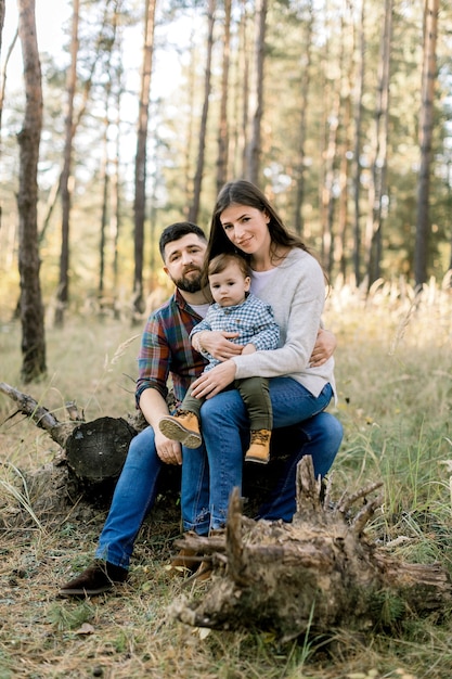 Ritratto all'aperto di famiglia caucasica alla moda felice in abiti casual, giovani genitori e figlio piccolo bambino carino, in una passeggiata nella bellissima pineta, seduto su un tronco di vecchio albero e guardando la fotocamera