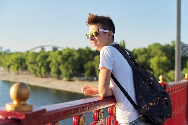 Ritratto all'aperto di giovane adolescente maschio sorridente con taglio di capelli alla moda che sta sul ponte sopra il fiume il giorno di estate soleggiato, spazio della copia