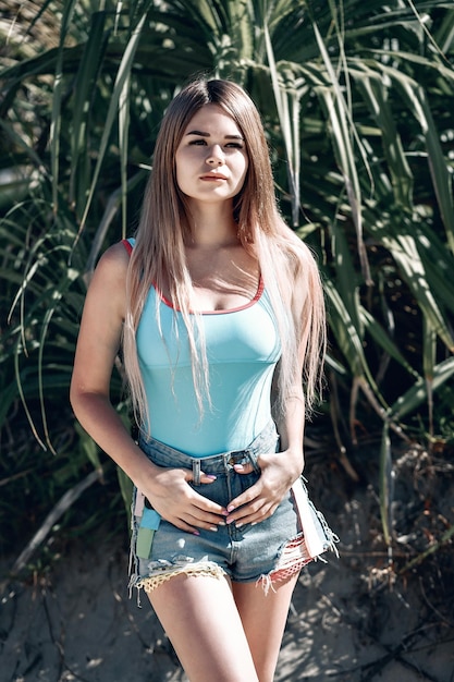 Ritratto all'aperto di giovane donna alla moda che indossa maglietta blu o top e pantaloncini di jeans blu in piedi sulla spiaggia vicino alle palme. Ragazza abbastanza sexy dei pantaloni a vita bassa. Tailandia