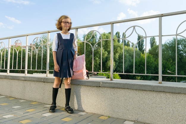 Ritratto all'aperto di piccola studentessa, ragazza con gli occhiali, uniforme con lo zaino