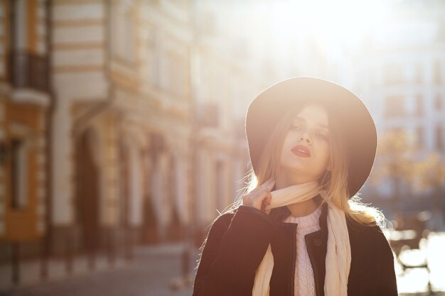 Ritratto all'aperto di seducente donna bionda che indossa cappello, sciarpa e cappotto, in posa sulla vecchia strada nel bagliore del sole. Spazio per il testo