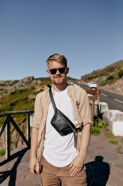 Ritratto all'aperto di un uomo europeo alla moda con i capelli biondi che indossa una camicia beige e una maglietta bianca in occhiali da sole che posano alla macchina fotografica alla luce del sole contro la strada pittoresca