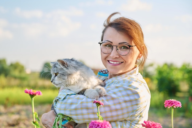 Ritratto all'aperto di una donna di mezza età con un gatto in braccio Felice guardando la fotocamera sorridente femmina matura con un soffice gatto grigio in campagna nella natura Amore sentimenti emozioni stile di vita animali persone