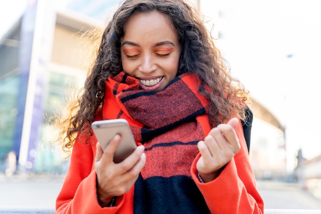 Ritratto all'aperto di una donna felice in cappotto arancione e sciarpa utilizzando un telefono cellulare e ridendo