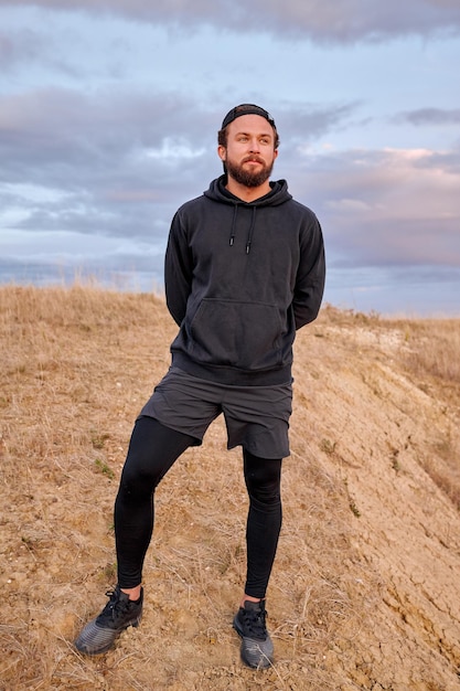 Ritratto all'aperto di uomo in abito sportivo nero in piedi nella natura sul campo, ragazzo caucasico fiducioso conduce uno stile di vita sano, impegnato nello sport, attività fisica. il maschio riposa dopo aver fatto jogging